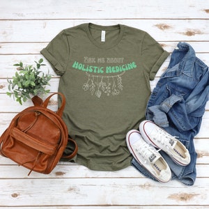 May include: Olive green t-shirt with the text "Ask Me About Holistic Medicine" in a mint green and white design. The shirt is styled with a brown backpack, white sneakers, and a denim jacket.