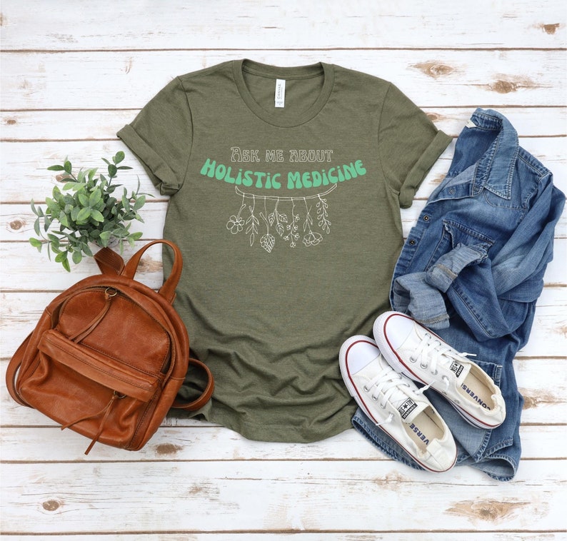 May include: Olive green t-shirt with the text "Ask Me About Holistic Medicine" in a mint green and white design. The shirt is styled with a brown backpack, white sneakers, and a denim jacket.