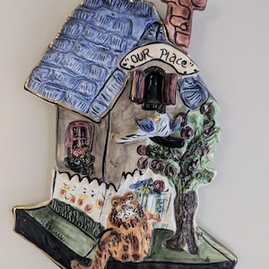 May include: A ceramic house-shaped wall hanging with a blue roof, chimney, and the text "Our Place". The artwork features a cat, a bird, and a tree with red fruit. The base is green with the artist's signature.