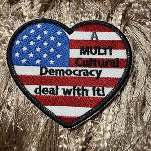 May include: Heart-shaped patch featuring a design inspired by the American flag. The patch is divided into two sections: a blue field with white stars and red and white stripes. Text reads "A Multicultural Democracy deal with it!"