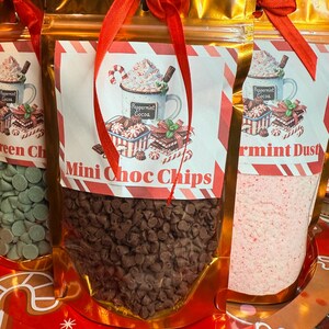 May include: Three gold-colored pouches tied with red ribbons, each containing different ingredients for hot cocoa. The center pouch is labeled "Mini Choc Chips" and contains small, dark brown chocolate chips. The other pouches contain green chocolate chips and pink peppermint dust.