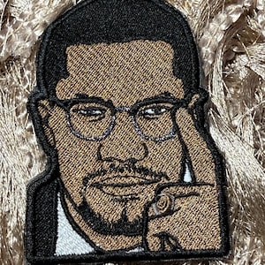May include: Embroidered patch featuring a portrait of a man with glasses, a beard, and a ring. The patch has a black border and is set against a textured background. The man is touching his glasses with his finger.