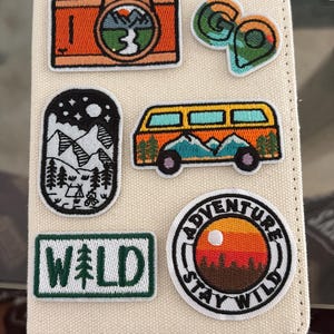 May include: A cream-colored fabric notebook cover decorated with six embroidered patches featuring outdoor themes, including an orange camera, a green map pin, a black and white mountain scene, an orange camper van, the word WILD, and a sunset circle.