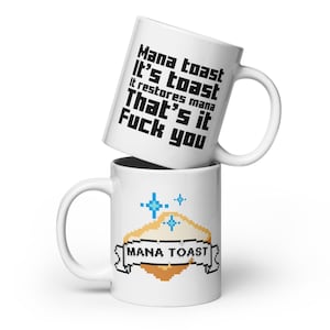 以下が含まれることがあります： 2つの白いセラミックマグ。上のマグには、黒いピクセルフォントで「Mana toast It's toast It restores mana That's it Fuck you」というテキストがあります。下のマグには、「Mana Toast」という言葉が書かれたピクセル化されたトーストのデザインが施されています。