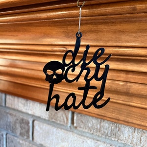 May include: Black metal ornament with a skull and the words "die cry hate" hanging from a wooden surface.