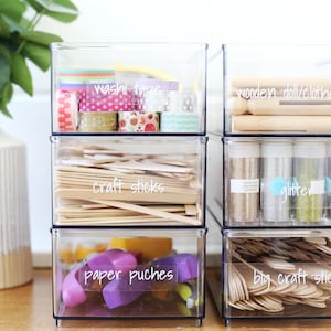May include: Clear plastic storage containers stacked on top of each other. The containers are labeled with white handwritten labels: "washi tape", "wooden doll/clothes pegs", "glitter", "craft sticks", "paper punches", and "big craft sticks".