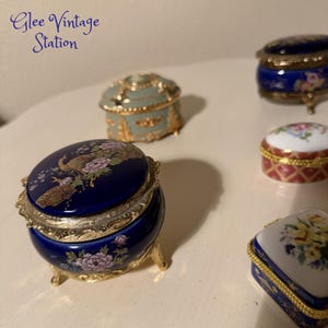 May include: Several decorative trinket boxes in various shapes and colors are displayed. The boxes feature floral and peacock designs, with gold-colored accents. The text "Glee Vintage Station" is visible in the upper left corner.