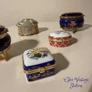 May include: A collection of small, decorative porcelain trinket boxes in various shapes and colors. The boxes feature floral designs, gold accents, and intricate details. One box is square with a floral lid, while others are round or oval.