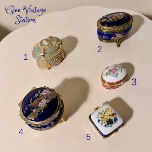 May include: A collection of five vintage porcelain trinket boxes. The boxes feature intricate designs, including floral patterns, a cameo, and a peacock. Colors include blue, gold, white, red, and green. The boxes are numbered 1 through 5.