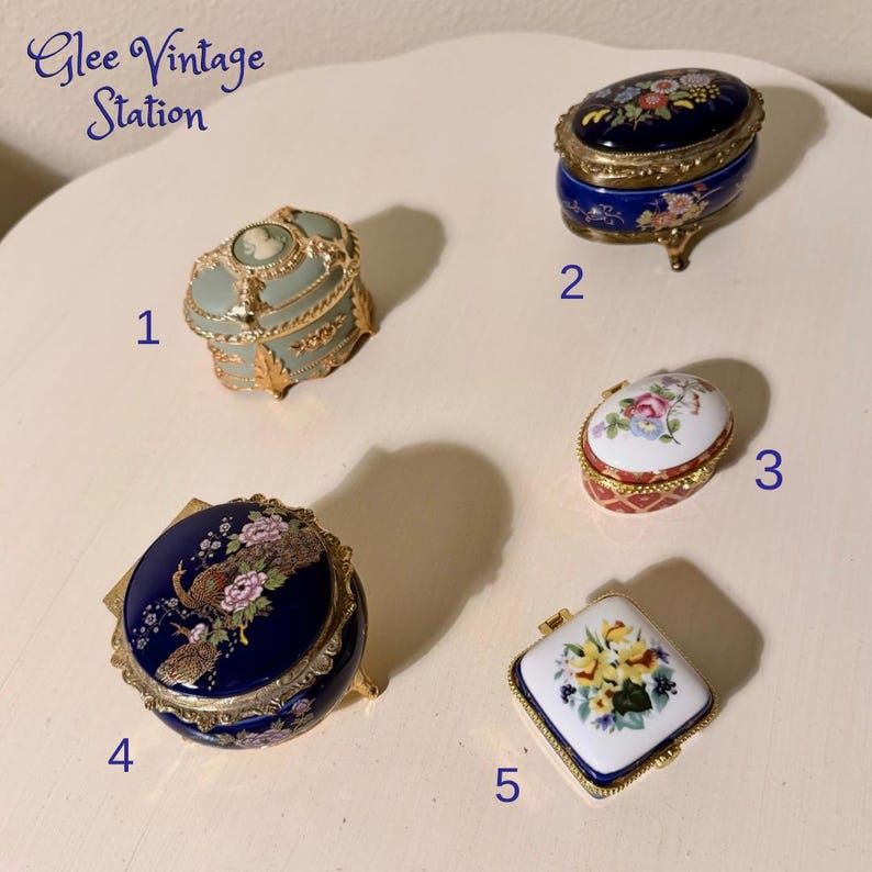 May include: A collection of five vintage porcelain trinket boxes. The boxes feature intricate designs, including floral patterns, a cameo, and a peacock. Colors include blue, gold, white, red, and green. The boxes are numbered 1 through 5.