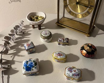 Könnte beinhalten: Eine Sammlung von sieben kleinen, dekorativen Boxen mit floralen und geometrischen Mustern. Die Boxen sind aus Porzellan und haben goldene Akzente. Die Boxen sind auf einer weißen Oberfläche mit einer Uhr und einer Schale mit Goldmünzen im Hintergrund angeordnet.