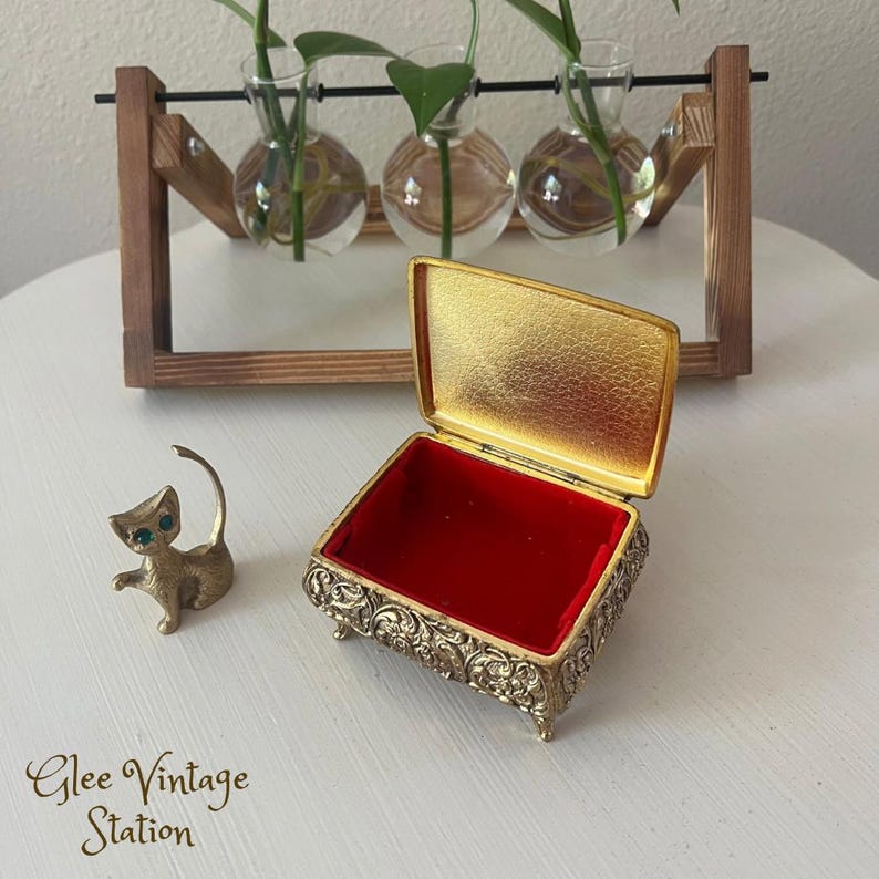 K&ouml;nnte beinhalten: Eine goldfarbene Schmuckschatulle mit einem verzierten Design und einem roten Samt-Innenraum. Die Schachtel ist ge&ouml;ffnet und steht auf einer wei&szlig;en Oberfl&auml;che. Eine kleine goldfarbene Katzenfigur mit gr&uuml;nen Augen befindet sich ebenfalls auf der Oberfl&auml;che.