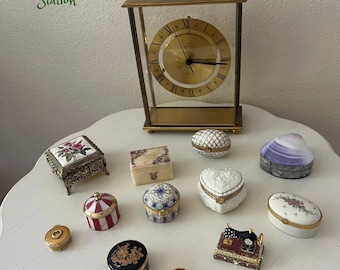 May include: A collection of vintage trinket boxes and a gold-toned clock on a white table. The boxes feature various shapes, colors, and designs, including floral patterns, stripes, and a sewing machine. The clock has a gold face and Roman numerals.