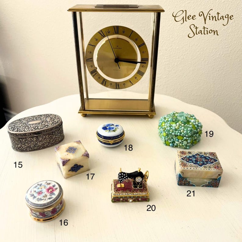 K&ouml;nnte beinhalten: Eine Sammlung von Vintage-Schmuckk&auml;stchen, darunter eine silberne ovale Schachtel mit einem verzierten Design, eine wei&szlig;e Schachtel mit blauen und goldenen floralen Akzenten, eine wei&szlig;e Schachtel mit einem N&auml;hmaschinen-Design, eine gr&uuml;ne Schachtel mit einem Perlen-Design, eine wei&szlig;e Schachtel mit einem floralen Design und eine wei&szlig;e Schachtel mit einem bunten Mosaik-Design. Die Schachteln sind auf einer wei&szlig;en Oberfl&auml;che mit einer goldenen Uhr im Hintergrund angeordnet. Der Text "Glee Vintage Station" ist in der oberen rechten Ecke des Bildes sichtbar.