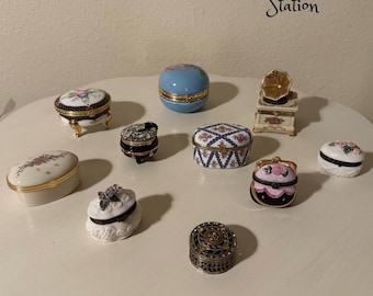 May include: A collection of decorative trinket boxes in various shapes and sizes. The boxes feature floral and geometric patterns, with gold accents. Some boxes are oval, round, or purse-shaped, and are made of porcelain or metal. The text "Glee Vintage Station" is visible.