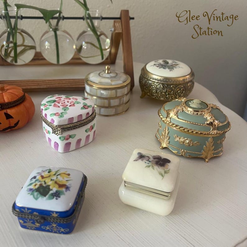 K&ouml;nnte beinhalten: Eine Sammlung von sechs Vintage-Schmuckk&auml;stchen. Die K&auml;stchen sind aus verschiedenen Materialien gefertigt, darunter Porzellan, Metall und Holz. Sie sind mit Blumenmustern, goldenen Akzenten und filigranen Designs verziert. Die K&auml;stchen sind auf einer wei&szlig;en Oberfl&auml;che angeordnet, im Hintergrund ist der Text "Glee Vintage Station" zu sehen.