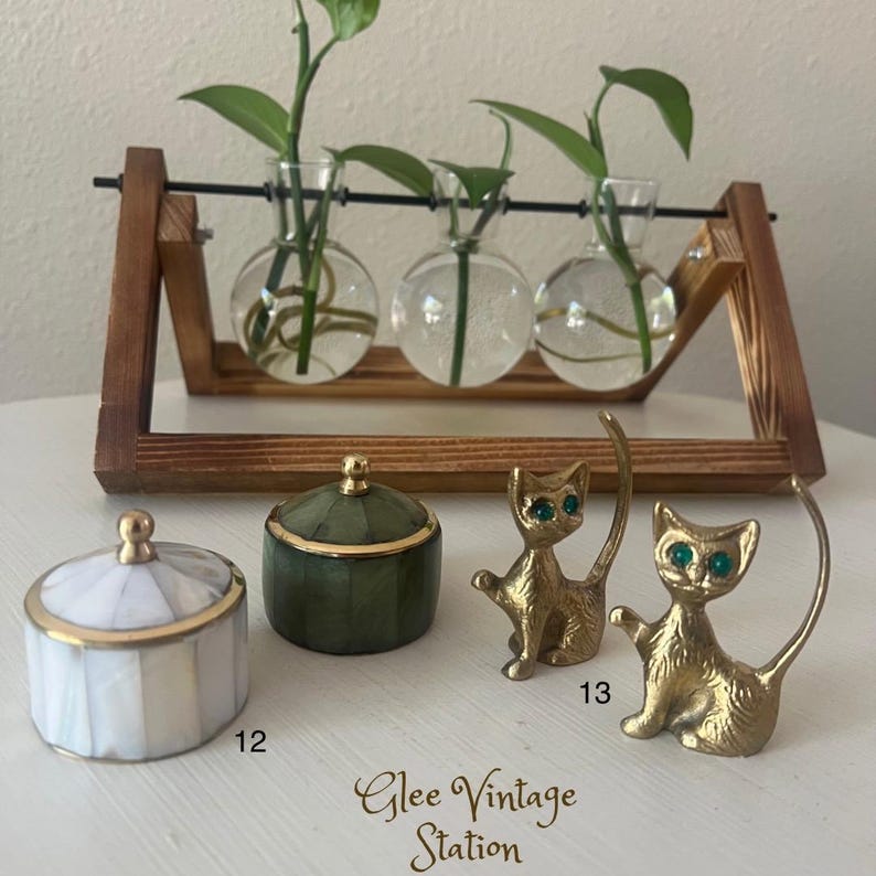 K&ouml;nnte beinhalten: Zwei kleine Dekorationsboxen, eine mit einem wei&szlig;en und goldenen Muschel-Design und die andere mit einem gr&uuml;nen und goldenen Design. Zwei goldene Katzenfiguren mit gr&uuml;nen Augen stehen auf einer Holzoberfl&auml;che. Der Text "Glee Vintage Station" ist unter den Figuren sichtbar.