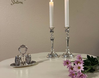 May include: Two silver candle holders with white candles lit, a silver salt and pepper shaker set with grape designs, and a bouquet of pink flowers on a white table.