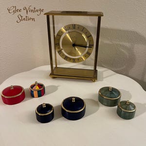 Könnte beinhalten: Eine goldfarbene Uhr mit römischen Ziffern auf dem Zifferblatt, die auf einer weißen Oberfläche mit fünf kleinen Dekorationsboxen steht. Die Boxen sind in verschiedenen Farben und Mustern, darunter Rot, Blau, Grün und Mehrfarbig.