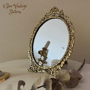 Könnte beinhalten: Ein ovaler Handspiegel in Goldton mit einem verzierten Rahmen mit floralem Muster. Der Spiegel steht auf einem Ständer und hat eine klare Reflexion.