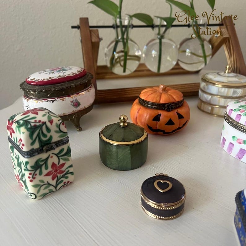 K&ouml;nnte beinhalten: Eine Sammlung kleiner, dekorativer Schmuckk&auml;stchen. Die K&auml;stchen sind aus Porzellan und haben verschiedene Designs, darunter einen K&uuml;rbis, ein Blumenmuster und eine gr&uuml;ne Schachtel mit goldenen Akzenten. Die K&auml;stchen sind auf einer wei&szlig;en Oberfl&auml;che angeordnet.