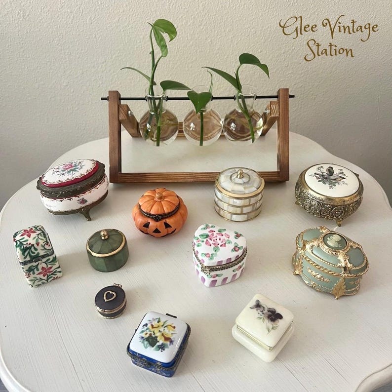 K&ouml;nnte beinhalten: Eine Sammlung von Vintage-Schmuckk&auml;stchen mit verschiedenen Designs und Farben, darunter eine K&uuml;rbis-f&ouml;rmige Schachtel, eine herzf&ouml;rmige Schachtel und eine runde Schachtel mit einem Blumenmuster. Die Schachteln sind auf einem wei&szlig;en Tisch mit einem Holzst&auml;nder angeordnet, der drei Glasvasen mit Pflanzen h&auml;lt.