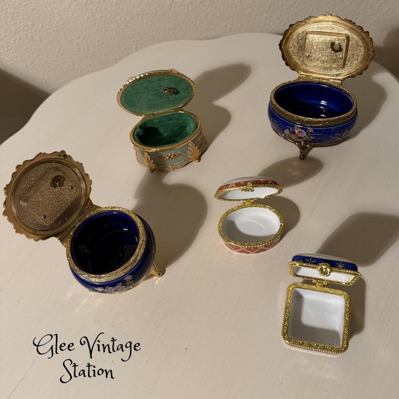 May include: A collection of vintage decorative trinket boxes. The boxes are in various shapes and sizes, with gold-toned metal frames and colorful enamel details. Some have hinged lids, revealing velvet or painted interiors.