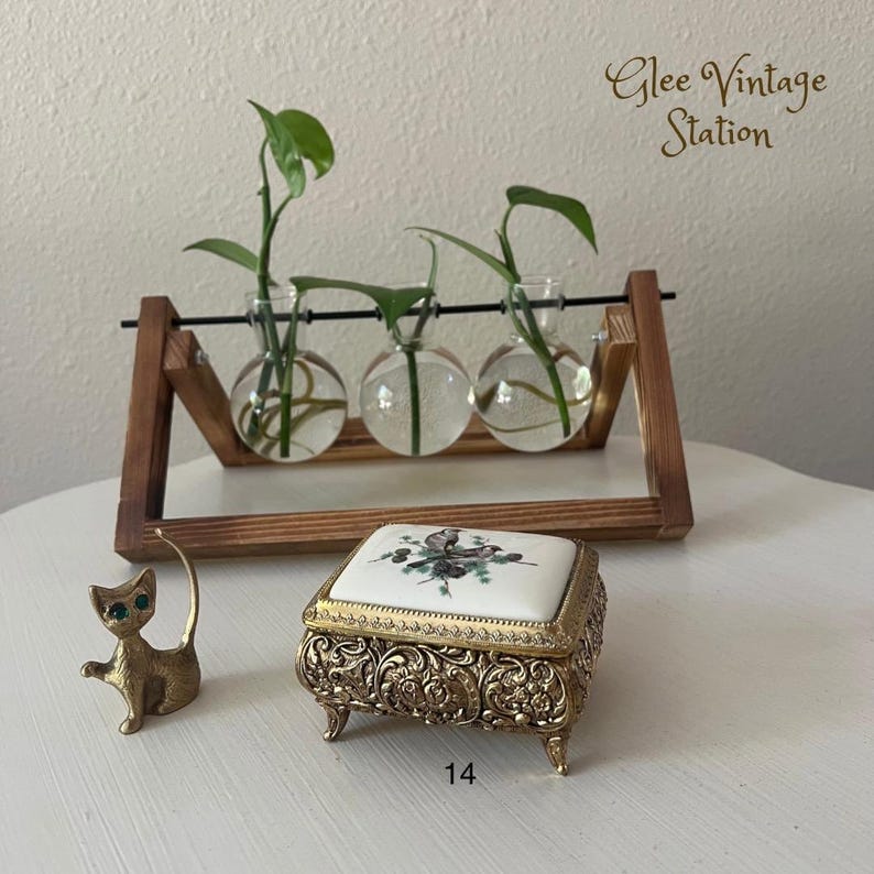 K&ouml;nnte beinhalten: Eine goldfarbene Schmuckschatulle mit einem wei&szlig;en Porzellan-Deckel, der mit einem Blumendesign verziert ist. Die Schatulle steht auf einem Holztisch mit einer kleinen goldenen Katzenfigur und einem Holz-Pflanzenst&auml;nder mit drei Glaskugeln, die Pflanzen enthalten.