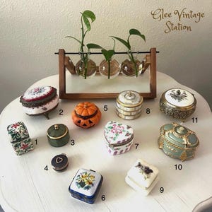 K&ouml;nnte beinhalten: Eine Sammlung von 11 Vintage-Schmuckk&auml;stchen mit verschiedenen Designs und Farben. Die K&auml;stchen sind auf einem wei&szlig;en Tisch mit einem h&ouml;lzernen Pflanzenst&auml;nder im Hintergrund angeordnet. Der Text "Glee Vintage Station" ist in der oberen rechten Ecke des Bildes sichtbar.