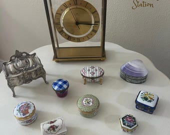May include: A collection of vintage decorative boxes and a brass clock on a white surface. The boxes feature floral and geometric designs, with a variety of shapes and colors. The clock has a gold face and Roman numerals. The text "Glee Vintage Station" is visible.