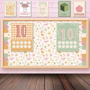 May include: A wooden framed noticeboard with two cards showing the number 10, surrounded by mushroom and leaf patterns. Above, are small framed prints with motivational quotes and geometric shapes. The background is a pink brick wall.