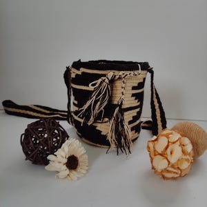 Puede incluir: Un bolso tipo cubo tejido y cilíndrico con un patrón geométrico en negro y beige, una correa larga a juego y dos borlas decorativas que cuelgan del cierre de cordón. El bolso está acompañado por una esfera de ratán marrón oscuro.