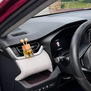 Może przedstawiać: Odświeżacz powietrza do samochodu z drewnianym klockiem i dwoma drewnianymi kulkami. Szklana butelka zawiera żółty płyn. Odświeżacz powietrza umieszczony jest na desce rozdzielczej samochodu.