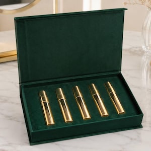 May include: A dark green velvet-lined presentation box containing five cylindrical gold-toned perfume atomizers arranged in a row. The box features a hinged lid that stands upright, showcasing the gold containers set against the deep green interior.