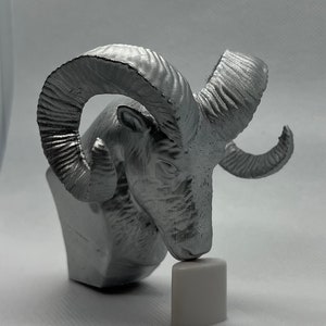 May include: A silver-colored ram head sculpture with large, curled horns. The sculpture is on a small, white base.