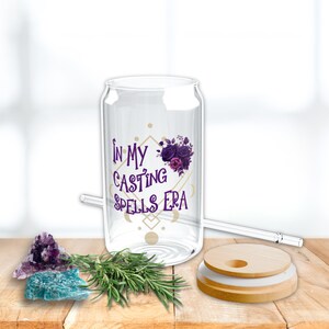 May include: A clear glass can with a lid and straw. The can has a purple floral design and the text "In My Casting Spells Era". The can is sitting on a wooden surface with a purple and blue crystal, a sprig of rosemary, and a wooden lid.