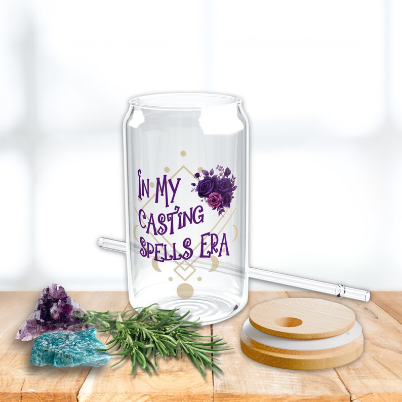 May include: A clear glass can with a lid and straw. The can has a purple floral design and the text "In My Casting Spells Era". The can is sitting on a wooden surface with a purple and blue crystal, a sprig of rosemary, and a wooden lid.