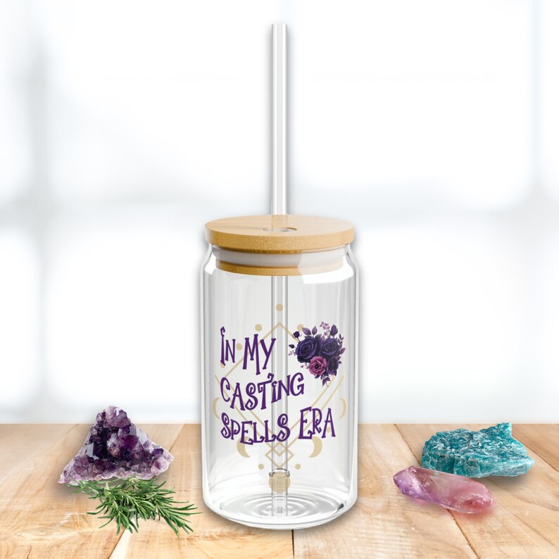 May include: A clear glass reusable water bottle with a bamboo lid and a straw. The bottle has a purple and gold design with the text "In My Casting Spells Era".