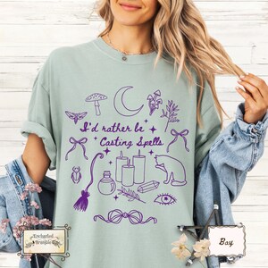May include: Sage green t-shirt with a purple graphic design featuring a crescent moon, mushrooms, candles, a cat, and the text "I'd rather be Casting Spells." The design includes stars, a potion bottle, and other mystical elements.