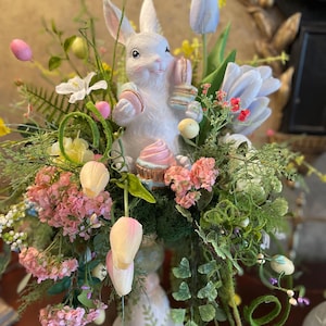 May include: A white ceramic bunny holding pastel macarons sits in a spring floral arrangement. The arrangement includes pink hydrangeas, yellow tulips, and green ferns. The arrangement is in a white ceramic pedestal.