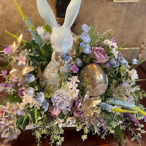 Può includere: Un centrotavola pasquale con un coniglietto in ceramica azzurra, lunghe orecchie, un fiocco dorato e un uovo decorativo. La composizione include fiori artificiali viola, blu e oro, creando un'esposizione festosa.