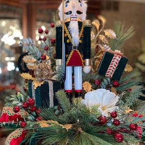 May include: A festive Christmas centerpiece featuring a traditional nutcracker soldier figurine. The arrangement includes green foliage, red berries, gold accents, and velvet gift boxes. The nutcracker is dressed in a black uniform with gold trim and a red skirt.