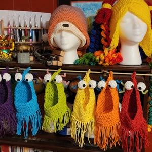 Może przedstawiać: Rząd siedmiu dzianych czapek w kolorach tęczy z frędzlami zwisającymi w dół. Każda czapka ma dwa duże wypukłe oczy. Czapki wiszą na drewnianym pręcie.