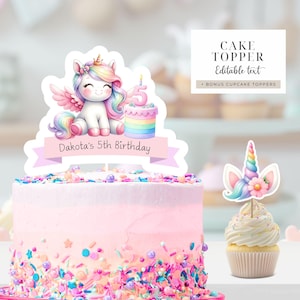 Puede incluir: Un topper de pastel de unicornio rosa y blanco con una melena y cola arcoíris, sosteniendo un pastel arcoíris con una vela encendida en la parte superior. El topper tiene una pancarta que dice "Dakota's 5th Birthday".
