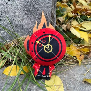 May include: A plush toy in the shape of a clock-faced character with a red and black design, yellow clock hands, and orange flames. The character is wearing a red suit with a black tie. The toy is surrounded by grass and yellow leaves.