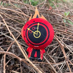 May include: A plush toy shaped like a clock with a red face, black center, and yellow clock hands. The toy has a small body with a red jacket and black pants. Orange flames are above the clock face. The toy is set against a background of brown twigs.