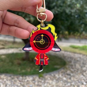 Könnte beinhalten: Ein farbenfroher Schlüsselanhänger mit einer Zeichentrickfigur mit einem Zifferblatt und einem rot-schwarzen Outfit. Der Schlüsselanhänger hat einen goldfarbenen Verschluss und Ring. Die Figur hat gelbe Haare und schwarze Ohren.