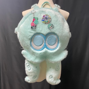 May include: A light mint green plush backpack shaped like a squid with two large circular eyes, dangling tentacles, and a top handle. The bag is decorated with a rectangular keychain, a circular holographic pin, and a small character pin.
