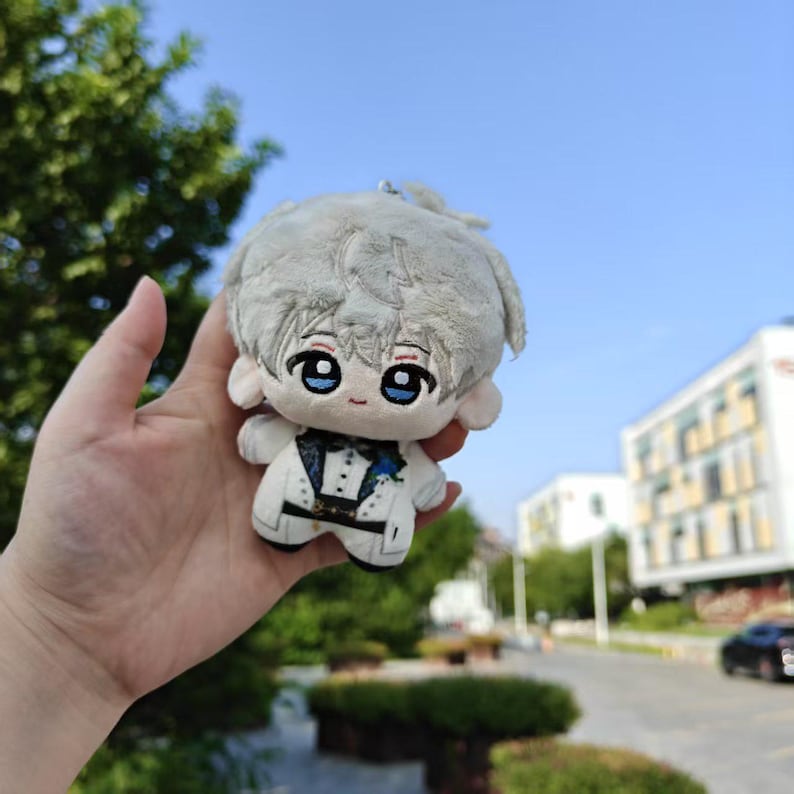 May include: A small plush doll with a light gray head and hair, large blue eyes, and a white suit with black accents. The doll is held in a hand against a backdrop of green trees and a blue sky.