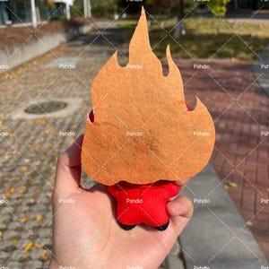May include: A plush toy shaped like a flame, with an orange felt flame and a red body. The toy is held in a hand, with the background showing a blurred outdoor scene.